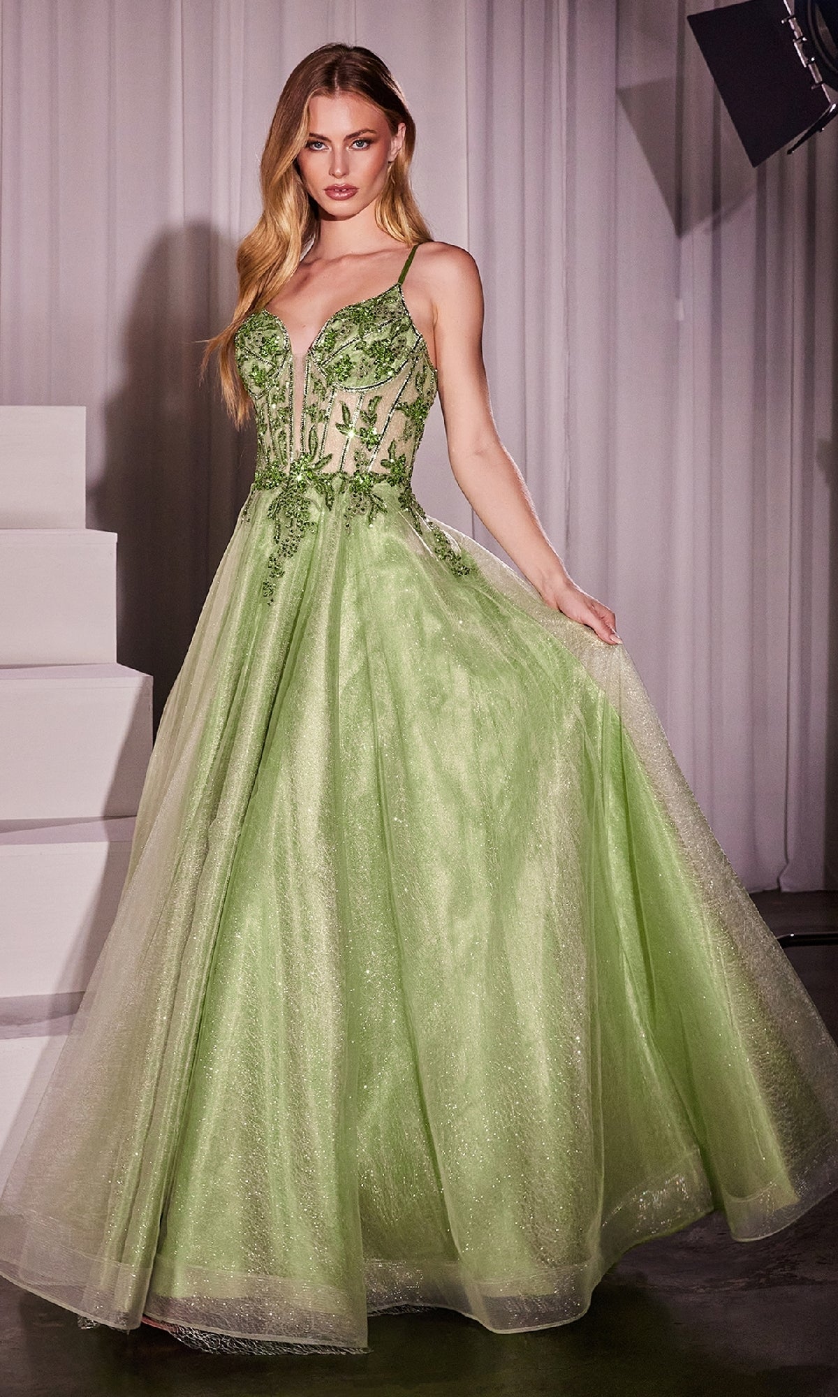 A woman stands indoors near white steps wearing the Sheer-Corset Long Prom Dress: Ladivine CC0881, a light green, floor-length gown featuring a fitted bodice with thin straps, floral appliqué detailing, and delicate beaded embroidery.